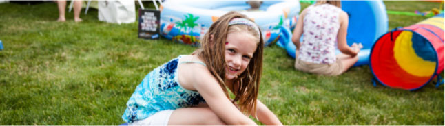 A girl in a blue patterned dress sits on the grass at a D10 family play area beside inflatable pools and a play tunnel.