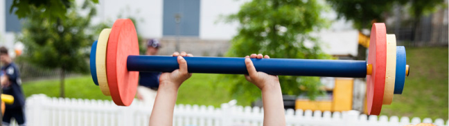 A child's hands press a colorful toy barbell overhead on a lawn beside a white picket fence.