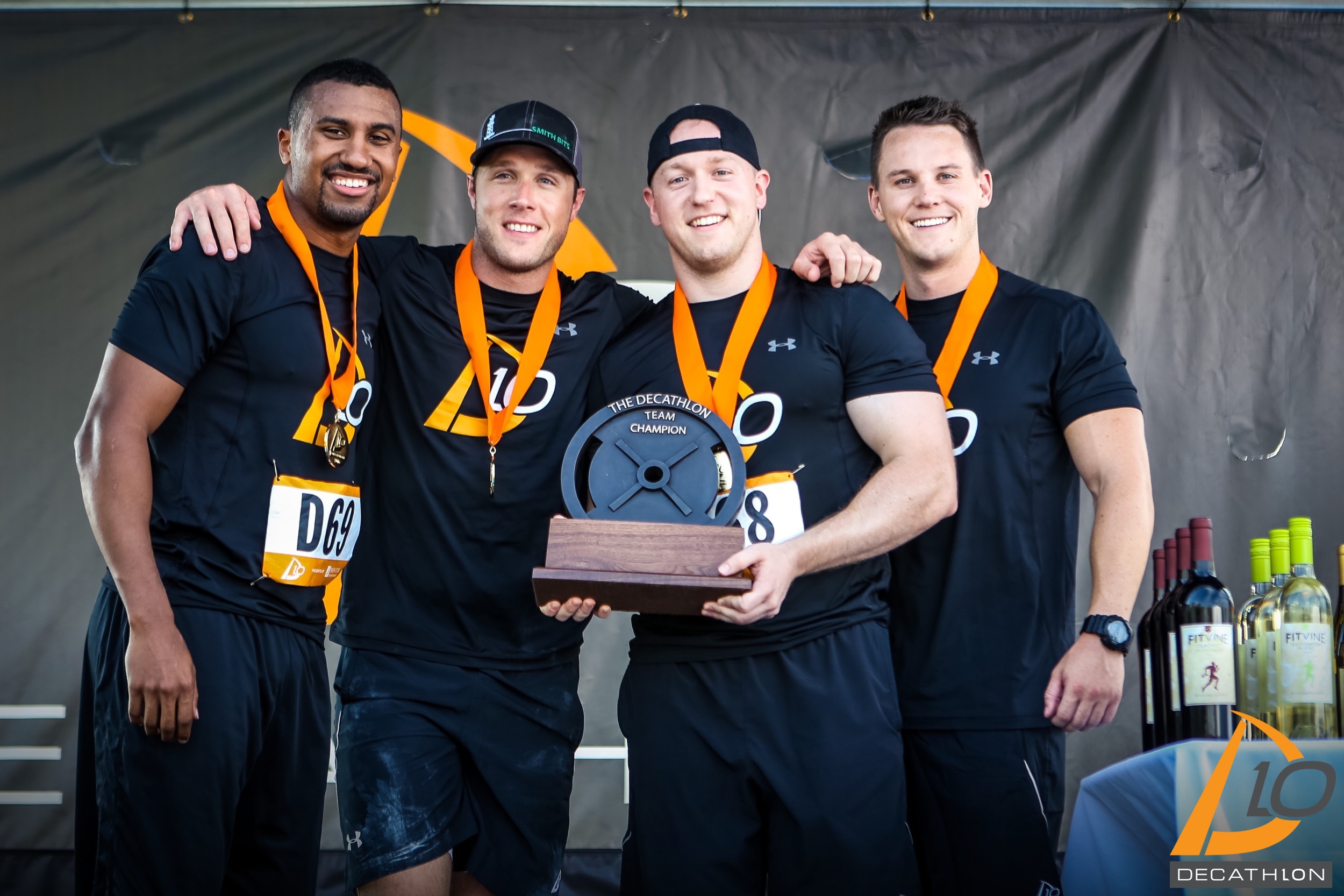 Four D10 teammates in black uniforms and orange medals pose with the Decathlon Team Champion trophy.