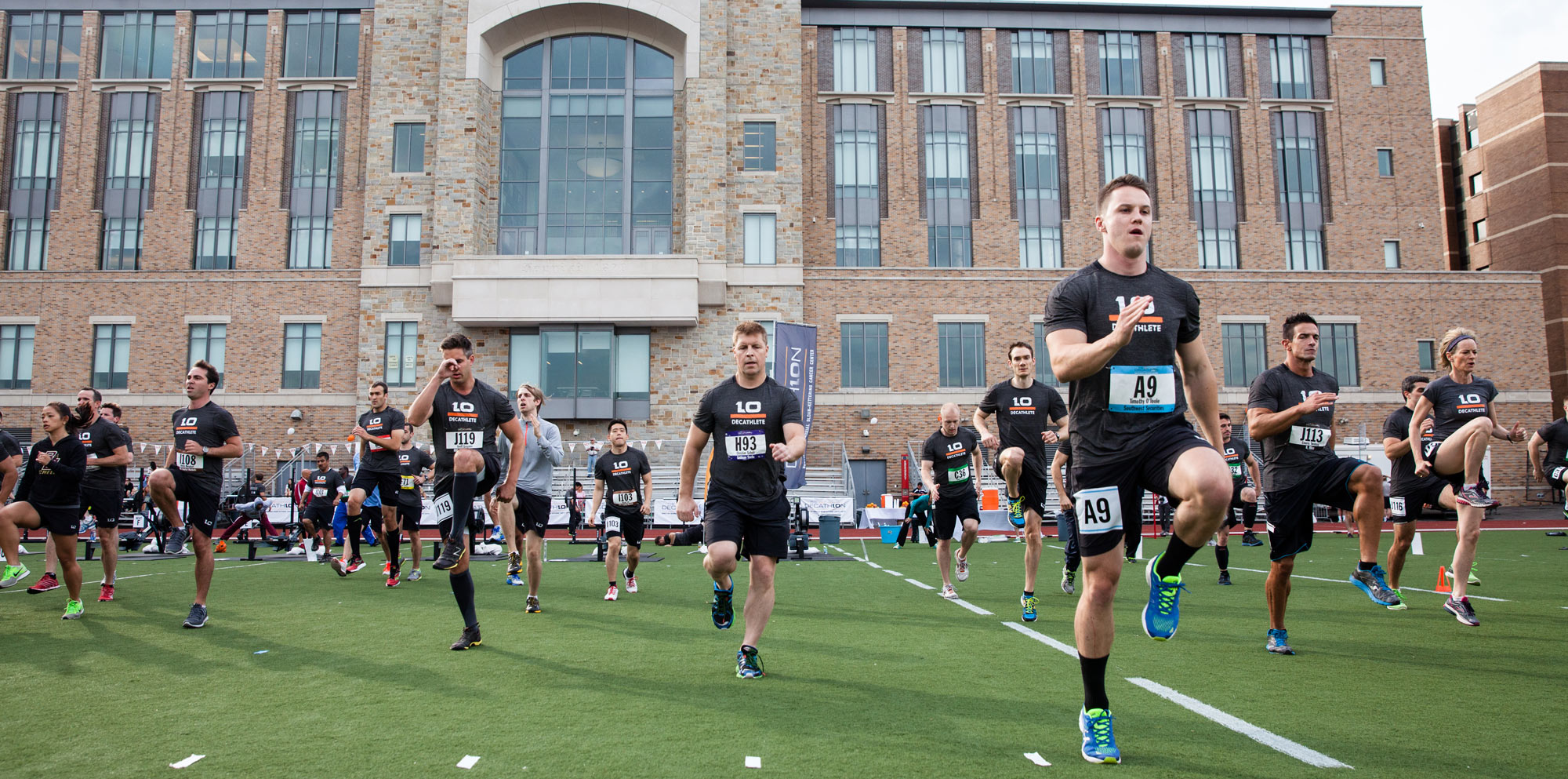 D10 athletes in black uniforms high-knee on the turf in front of a brick campus building during a team warmup.